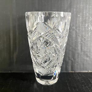 May include: Clear crystal vase with intricate geometric and starburst patterns. The vase has a wide mouth and a slightly tapered body. The cut crystal design reflects light, creating a sparkling effect. A decorative piece for home decor.