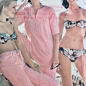 May include: Three women wearing swimsuits. Two women are wearing pink pants and a floral print bikini top. The third woman is wearing a floral print bikini top and bottom. The women are standing on a beach.