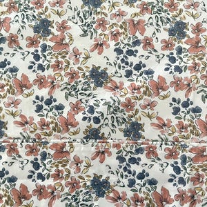 Op de afbeelding: Bloemenstof met een herhalend patroon van roze, blauwe en groene bloemen op een witte achtergrond. De bloemen zijn gestileerd en dicht op elkaar geplaatst, wat een dichte, getextureerde uitstraling creëert. De stof lijkt katoen of een vergelijkbaar materiaal te zijn.