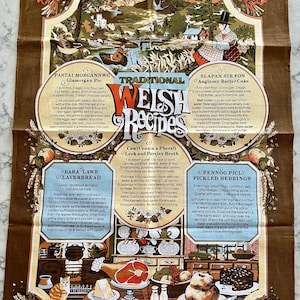 May include: A brown tea towel with a colorful illustration of a Welsh landscape and traditional Welsh recipes. The recipes include Glamorgan Pie, Bara Lawr Laverbread, Cawl Cennin a Phersli Leek and Parsley Broth, Slapan Sir Fon Anglesey Batter Cake, and Pennog Piclo Pickled Herrings.