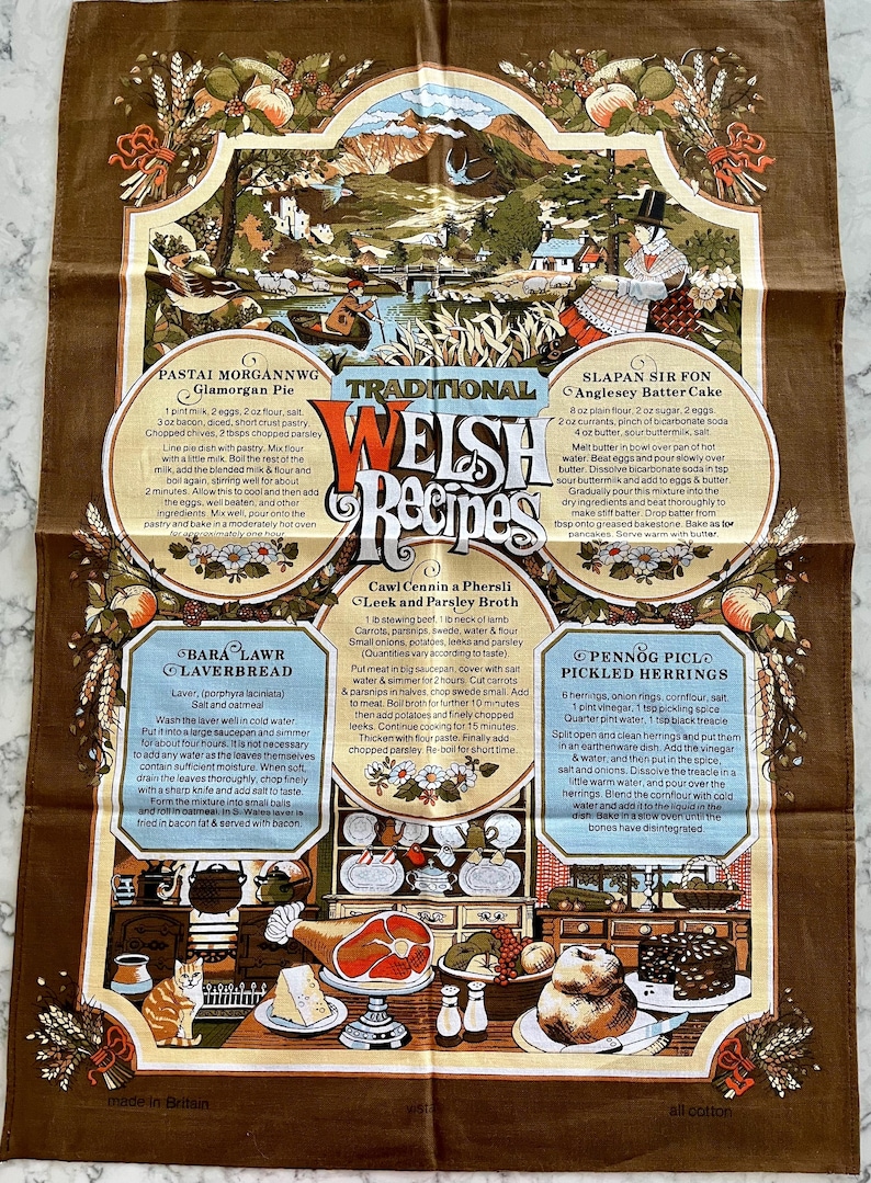May include: A brown tea towel with a colorful illustration of a Welsh landscape and traditional Welsh recipes. The recipes include Glamorgan Pie, Bara Lawr Laverbread, Cawl Cennin a Phersli Leek and Parsley Broth, Slapan Sir Fon Anglesey Batter Cake, and Pennog Piclo Pickled Herrings.