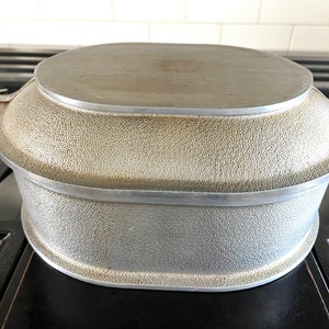 May include: A vintage silver-toned metal cooking pot with a textured surface and a matching lid. The pot has a rounded base and a flat, oval-shaped lid. The handles are decorative. The pot is sitting on a black stovetop.