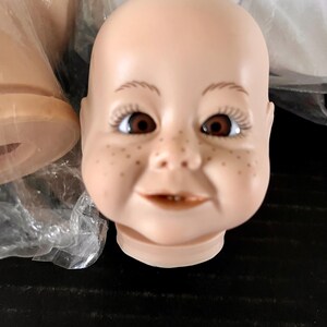 May include: A close-up of a porcelain doll head with brown eyes, a smiling mouth, and freckles. The head is unpainted and ready for customisation.