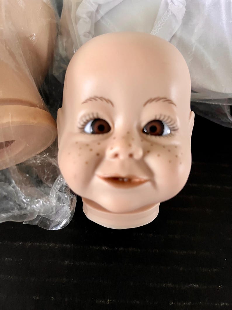 May include: A close-up of a porcelain doll head with brown eyes, a smiling mouth, and freckles. The head is unpainted and ready for customisation.