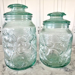 Peut inclure: Deux bocaux en verre vintage avec des couvercles verts et des motifs de fruits en relief. Les bocaux sont en verre transparent avec une finition givrée.
