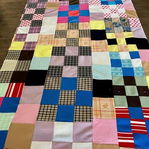 May include: A patchwork quilt with a variety of fabric squares in various colors and patterns. The quilt features squares of pink, blue, brown, and white fabrics, some with polka dots, plaid, and floral designs. The quilt is laid out on a wooden surface.