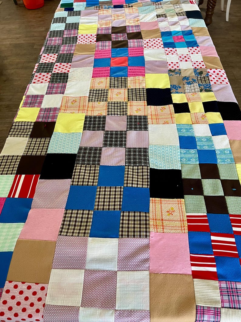 May include: A patchwork quilt with a variety of fabric squares in various colors and patterns. The quilt features squares of pink, blue, brown, and white fabrics, some with polka dots, plaid, and floral designs. The quilt is laid out on a wooden surface.