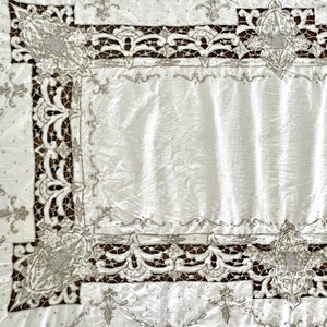 May include: White linen tablecloth with a delicate lace border and embroidered floral motifs. The tablecloth has a subtle sheen and a classic design.