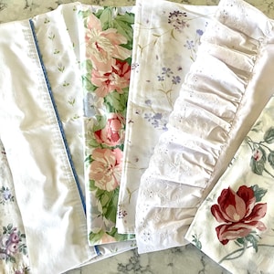 May include: Assortment of vintage floral bed sheets. The sheets feature various patterns, including large pink roses, small purple flowers, and white sheets with blue trim. Some sheets have eyelet lace and ruffled edges.