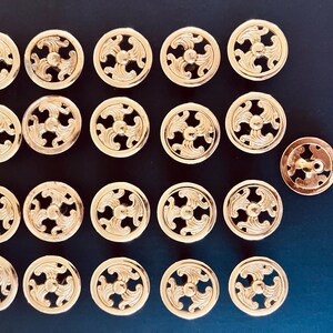 May include: A collection of twenty-one circular gold-toned metal buttons, each featuring an intricate openwork design with three symmetrical curved leaf-like motifs radiating from a central point, arranged in a grid pattern on a dark surface.