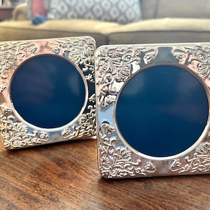 May include: Two square silver-toned picture frames featuring intricate embossed floral and leaf patterns along the borders. Each frame has a circular center opening currently filled with a solid dark blue insert, creating a high-contrast look.