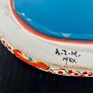 May include: Hand-painted ceramic plate with a white rim, red and orange floral accents, and a turquoise center. The plate has a decorative border with red dots. The text "A.J.M., MEX" is handwritten on the side.