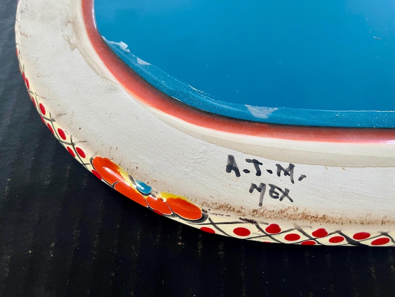 May include: Hand-painted ceramic plate with a white rim, red and orange floral accents, and a turquoise center. The plate has a decorative border with red dots. The text "A.J.M., MEX" is handwritten on the side.