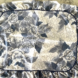 May include: A white and gray floral patterned pillowcase with a blue trim. The pillowcase features large, stylized flowers and leaves in shades of gray and blue. The edges are ruffled.