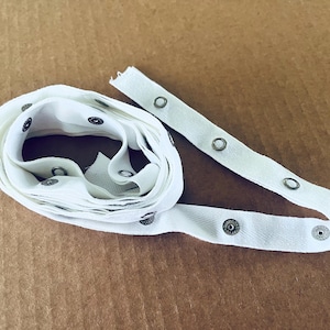 May include: A white fabric tape featuring a series of evenly spaced metal snap fasteners. The tape is coiled into a circular shape with one end extending outward, displaying the silver-toned circular snaps attached to the textile strip.