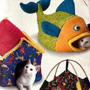 Puede incluir: Tres camas para gatos de varias formas y colores. Una es una cama con forma de pez con tela naranja y verde, y un gato blanco asomando. Otra es una cama con forma de casa con techo rojo y paredes azules, con un gato blanco dentro. La tercera es una cama con forma de tienda.