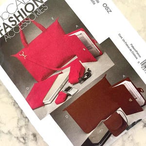May include: A McCall's Fashion Accessories sewing pattern envelope featuring illustrations of a tote bag, a laptop sleeve, a small pouch, and a wallet in both bright pink and dark brown fabric options, with a barcode and text M6091/OSZ.