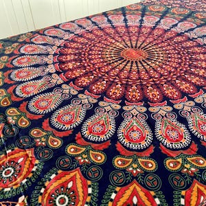 Puede incluir: Un gran tapiz colorido con un diseño de mandala. El tapiz presenta un patrón circular central en tonos de rojo, naranja y dorado, rodeado de intrincados motivos tipo paisley sobre un fondo azul marino. El diseño general es simétrico.