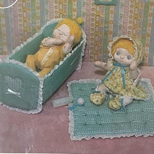 May include: A handmade doll set featuring a baby doll in a crib and a doll on a blanket. The crib and blanket are teal with white lace trim. The dolls have yellow hair and wear yellow and floral outfits.