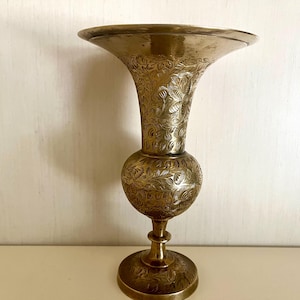 May include: A brass-colored vase with an ornate floral pattern. The vase has a flared top, a rounded midsection, and a pedestal base. The intricate design covers the entire surface of the vase.