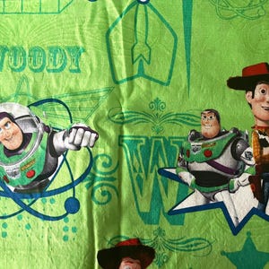 May include: Green fabric featuring characters from the film Toy Story. Buzz Lightyear is in a spacesuit, and Woody wears a cowboy hat. The word "WOODY" is printed in green on the fabric.