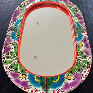 May include: Oval-shaped decorative mirror with a vibrant floral border. The border features purple, blue, green, and yellow flowers on a white background, with red accents. The mirror has a bright orange frame.