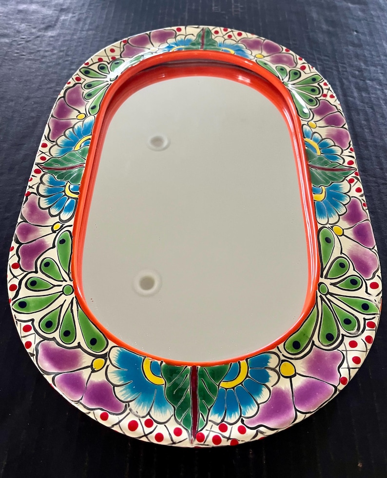 May include: Oval-shaped decorative mirror with a vibrant floral border. The border features purple, blue, green, and yellow flowers on a white background, with red accents. The mirror has a bright orange frame.