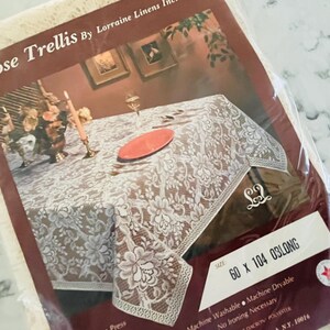 May include: A white lace tablecloth in plastic packaging labeled Rose Trellis by Lorraine Linens Inc. The packaging features a printed image of a table set with a red plate and candle. The label specifies a size of 60 x 104 inches for the tablecloth.