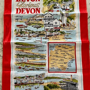 May include: A red tea towel with a white design featuring a map of Devon, England, with illustrations of various towns and cities, including Ilfracombe, Cockington Forge, Exeter, Torquay, Paignton, Brixham, Teignmouth, Westward Ho!, Clovelly, and The Hoe Plymouth. The text "Devon Glorious Devon" is printed at the top of the towel.
