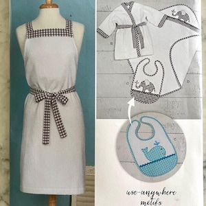 Puede incluir: Un delantal blanco con ribete a cuadros blanco y marrón y una cinta a juego. El delantal se muestra con una bata blanca y un babero blanco con un diseño de elefante bordado. El texto "use-anywhere motifs" está impreso debajo del babero.