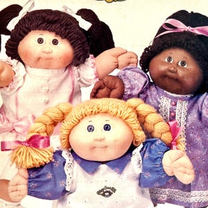 Puede incluir: Se muestran tres muñecas Cabbage Patch Kids, cada una con peinados y atuendos únicos. Una muñeca tiene cabello oscuro en coletas con lazos blancos, otra tiene coletas rubias con lazos rosas y la tercera tiene cabello negro y un lazo rosa. Las muñecas visten vestidos.