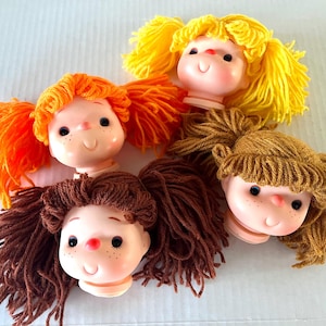 May include: Four vintage doll heads with yarn hair in orange, yellow, and brown. The doll heads have painted facial features, including black eyes, freckles, and a red nose. The dolls have a smiling expression.