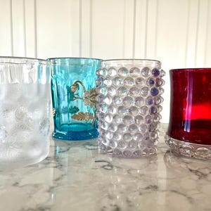 May include: Four decorative drinking glasses of varying designs. The first is clear with a floral pattern, the second is turquoise with gold accents, the third is clear with bubble-like texture, and the fourth is ruby red.