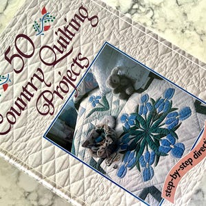 Pode incluir: Um livro intitulado "50 Country Quilting Projects" com uma capa acolchoada branca e uma foto de um edredom azul e verde com um design floral. O texto na capa diz "step-by-step directions!"