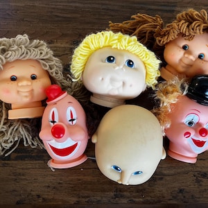 Pode incluir: Uma coleção de cabeças de bonecas vintage com vários penteados e expressões faciais. Inclui um palhaço, uma cabeça careca e bonecas com cabelo de fio loiro e castanho. As cabeças de boneca de plástico mostram sinais de desgaste.