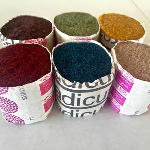 May include: Six skeins of yarn in various colors, including burgundy, teal, olive green, and mustard yellow. The yarn is wrapped in paper with printed text and a pink label. The image shows a close-up of the yarn, highlighting the texture and color variations.