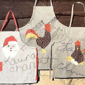 May include: Three handmade aprons with unique designs. One features Santa Claus, another a rooster, and the third a hen. Each apron is made of gray and white striped fabric and has red accents. The aprons have handwritten text.