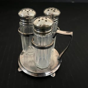 以下が含まれることがあります： 銀製の塩とコショウのセット。ガラス製のシェーカーが3つ、銀製のスタンドの上に置かれています。シェーカーは銀製の蓋が付いており、透明なガラス製です。