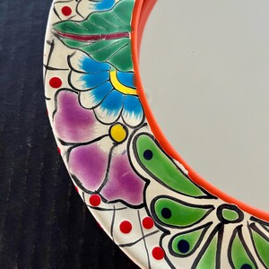 May include: A round mirror with a vibrant, hand-painted floral design. The mirror's frame is a bright red, and the decorative border features flowers in shades of purple, blue, and green, with red and yellow accents. The mirror's surface is reflective.