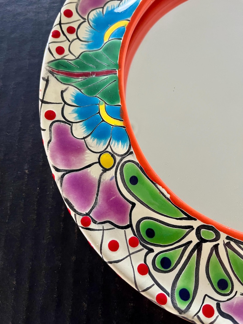 May include: A round mirror with a vibrant, hand-painted floral design. The mirror's frame is a bright red, and the decorative border features flowers in shades of purple, blue, and green, with red and yellow accents. The mirror's surface is reflective.