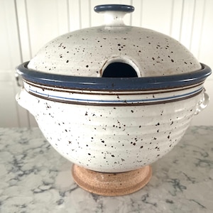 May include: A speckled white ceramic soup tureen with a lid and a round opening. The tureen has a blue rim and brown accents. It sits on a round, light brown base. The tureen has two handles on the sides.