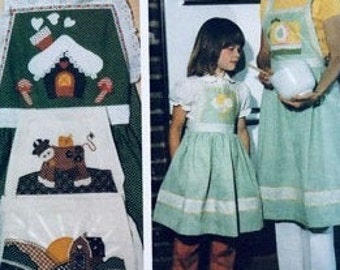 May include: Two green and white aprons with embroidered designs. The larger apron has a gingerbread house, candy canes, a cat, a deer, and a sunrise. The smaller apron has a daisy.