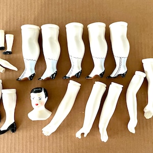 May include: A collection of vintage doll parts, including white porcelain legs with black and red heeled shoes, arms, and a doll head with painted features. The parts are arranged on a brown surface.