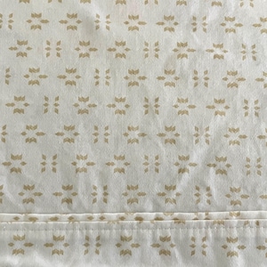 May include: Cream-colored fabric with a repeating pattern of gold-colored floral and geometric shapes. The design is evenly spaced across the material, with a stitched seam at the bottom. The fabric appears to be slightly textured.