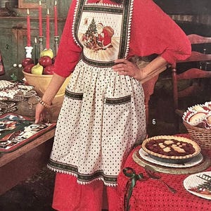 May include: A woman wearing a Christmas apron with a Santa Claus design. The apron is white with black polka dots and green trim. A table is set with a pie, placemats, and a basket of rolls. Red candles and apples are also visible.