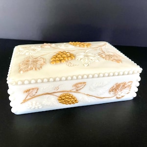 Peut inclure: Boîte à bijoux rectangulaire blanche avec couvercle, ornée de motifs de raisins et de feuilles en relief dorés et blancs. La boîte a une bordure perlée et un bord festonné, pour un style vintage.