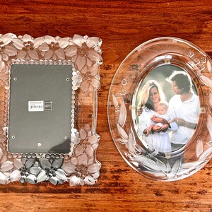May include: A rectangular glass picture frame with a frosted floral border and an oval glass picture frame with etched leaf patterns. The oval frame contains a photograph of two adults holding a baby. Both frames feature clear glass elements.