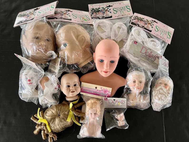 May include: An assortment of porcelain doll heads and parts for crafting. The heads are in various sizes and styles, including a baby, a young girl, an older woman, and a Santa Claus. The parts include a wig, a bust, and a pair of eyes. The packaging includes the text "Doll & Co. Everything for the Doll Maker".