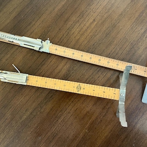 Può includere: Due strumenti di misurazione vintage in legno e metallo, probabilmente per la lavorazione del legno. Ogni strumento presenta un righello in legno con segni in centimetri e un componente metallico scorrevole. Gli strumenti sono su una superficie di legno scuro.
