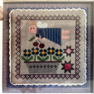 Peut inclure: Une broderie au point de croix encadrée représentant un ange à la peau claire avec une auréole et des ailes blanches, vêtu d'une robe bleue. L'ange tient deux cerises rouges au-dessus d'une jardinière grise remplie de fleurs bleues.